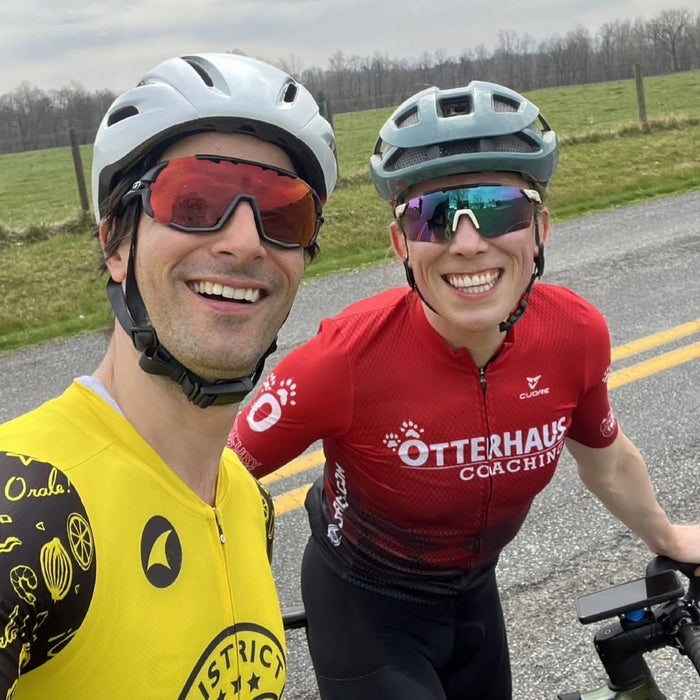Two cyclists one wearing Otterhaus Coaching jersey and one in yellow on a road with green fields and trees in the background.