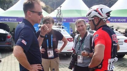 Two people in cycling gear talking to two other people, with a backdrop of cars and event banners.