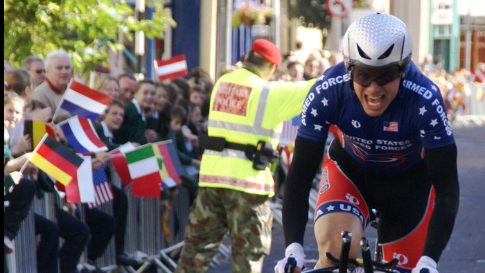Cyclist in a racing uniform riding a bike on a street with spectators and flags.