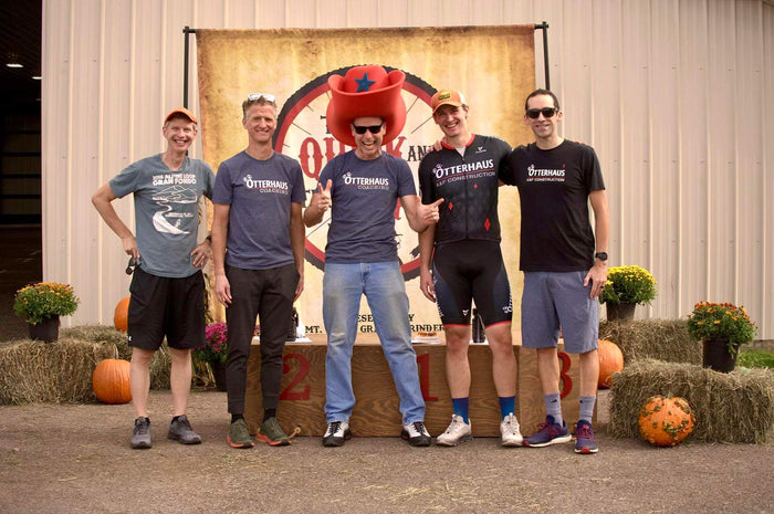 Five cyclists with Otterhaus Coaching shirts posing together in front of a large banner with pumpkins and hay bales.