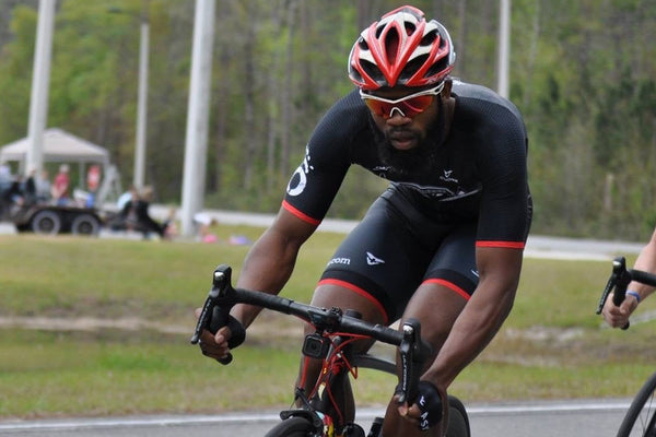 Cyclist in a black and red outfit with a helmet and sunglasses riding a bike on a road.