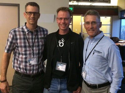 Three men standing together in an office setting, wearing lanyards with badges.