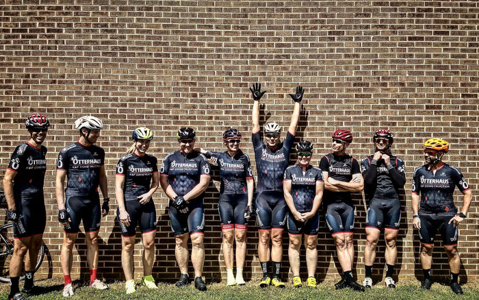Group of cyclists in matching Otterhaus uniforms standing against a brick wall.