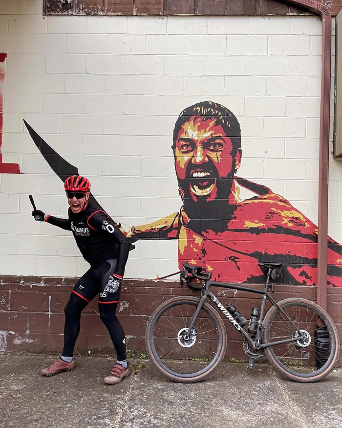 Cyclist posing in front of a bicycle and mural of a spartan with a large axe on a brick wall.