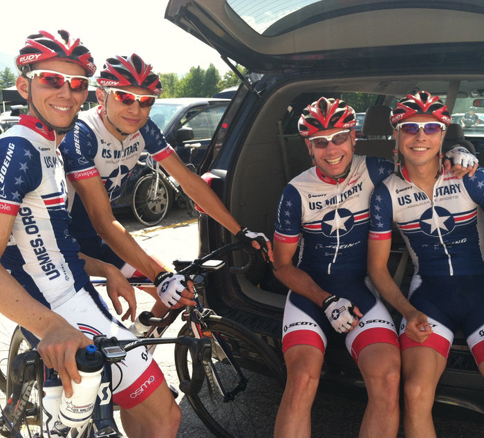 Four cyclists in US Military cycling uniforms sitting in the back of a car with bicycles.